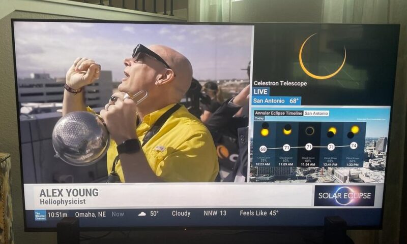 View of a TV screen with a man on it with yellow shirt wearing eclipse glasses and holding a colander looking up at the sun.