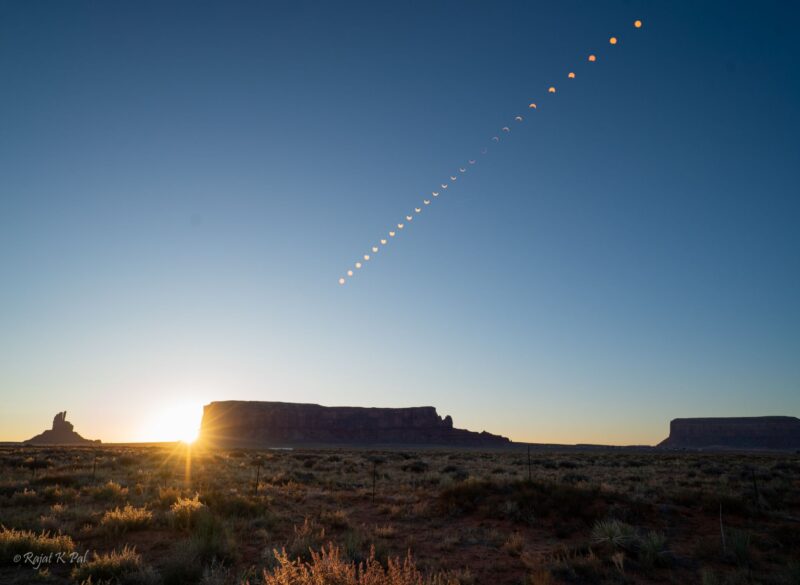 26 shapes for the eclipse on a blue sky. There are some mountains under the eclipse shapes.