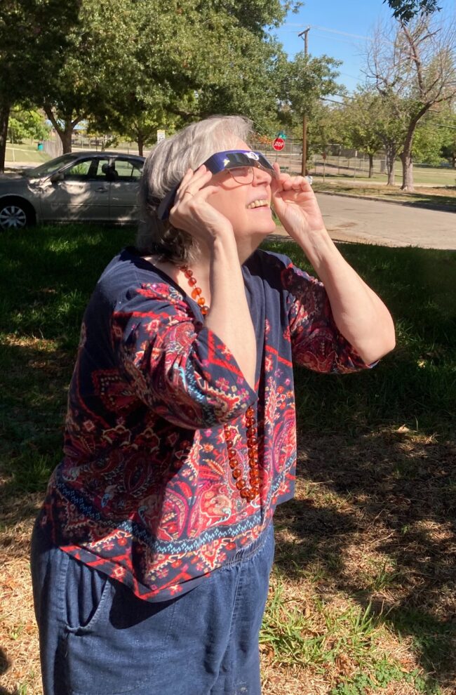 A gray-haired woman holds up eclipse glasses to her face and peers up as she stands in sunshine in a yard.