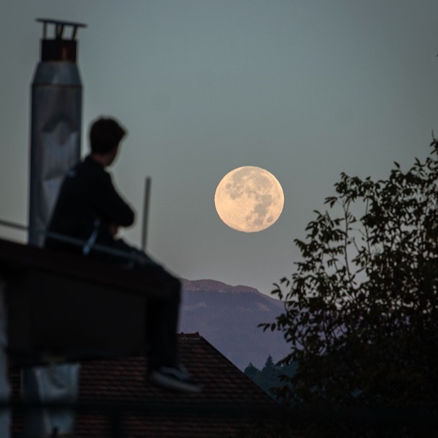 Silhouette of a sitting man watching the large, white full moon rise over a distant mountain.