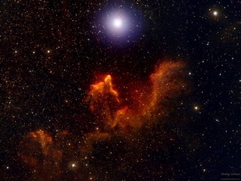 Very irregular, elongated orange cloud near large, bright star, in star field.