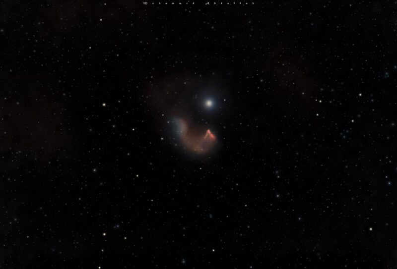 Foggy reddish C-shaped nebula near a bright star, with faint background stars.