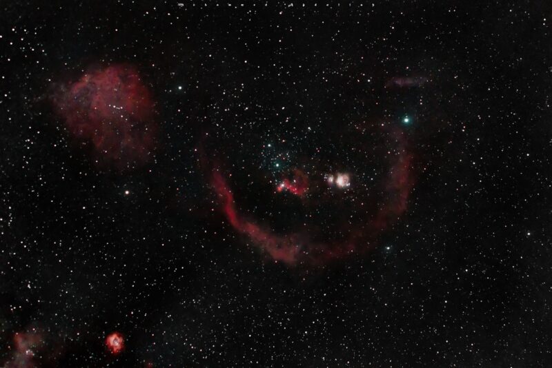 A dozen irregular reddish clouds, including one large semicircle, behind a multitude of foreground stars.