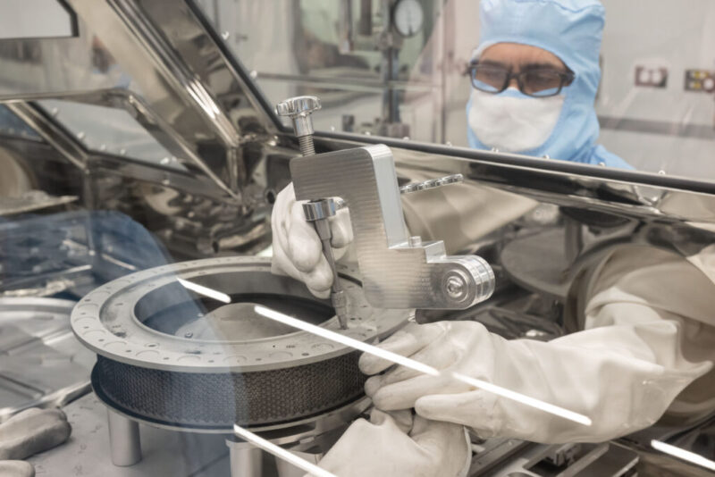 Scientist garbed in clean-room suit and heavy gloves working on a cylindrical metal container.