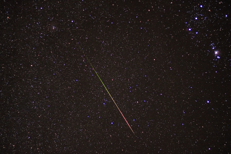 Bright colorful meteor streaking across a starry sky with some of the constellation Orion above it.