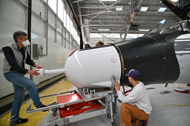 Three men installing a large white bullet-shaped nose cone on an airplane.