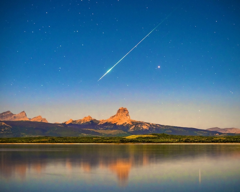 Long bright streak in starry blue sky above a lake with distant mountains.