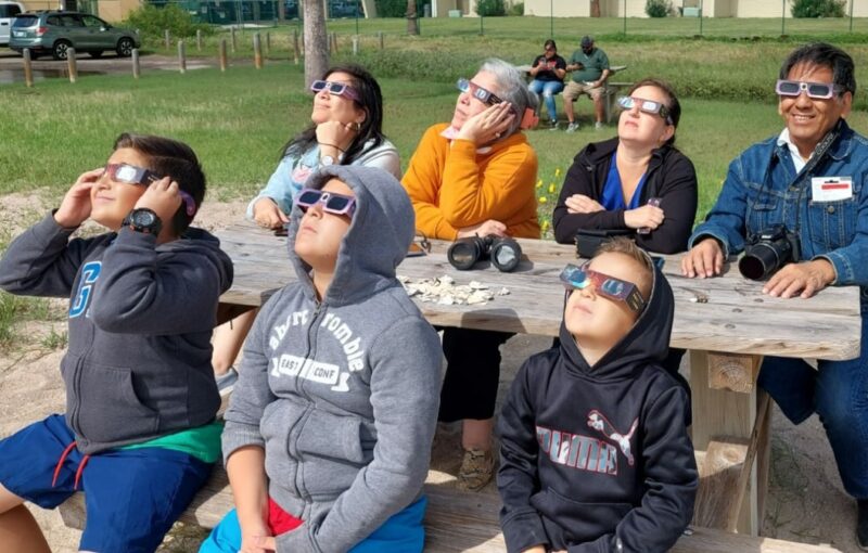 A group of adults and children sit at a picnic table, wearing eclipse glasses and looking up.
