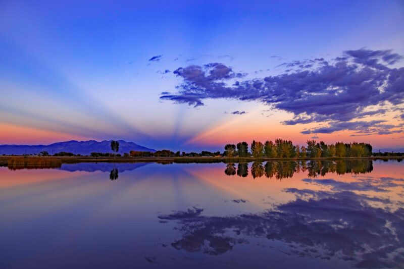 Blue rays radiating from an orange and blue sunset, with distant mountain, all reflected in calm lake.