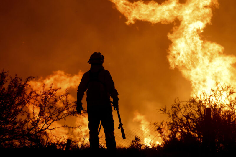 Silhouette of lone fireman standing in front of a bank of huge, leaping flames.