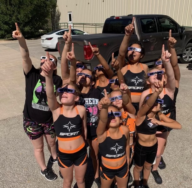 A group of young girls in cheer outfits and eclipse glasses, looking and pointing to the sky.