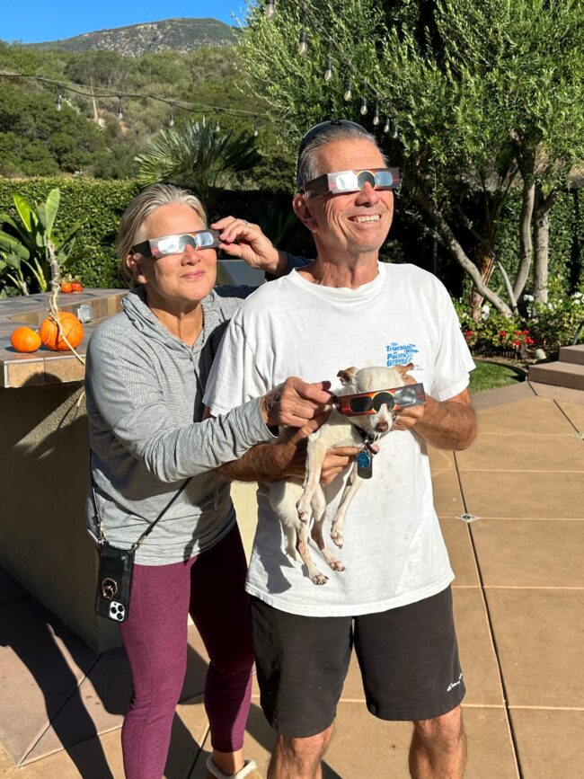 Two people holding a small dog, all wearing eclipse glasses.