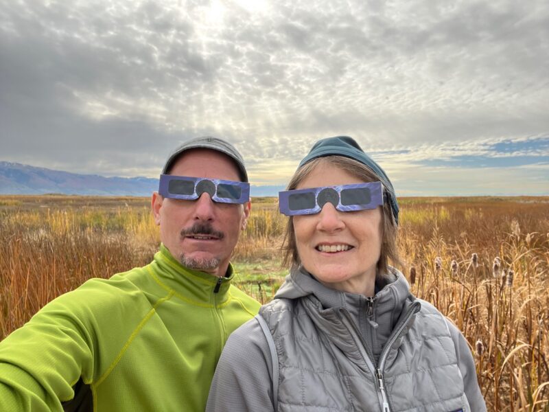 Man and woman, in an open field, wearing eclipse glasses. The sky looks cloudy and grey, but there are some areas without clouds.