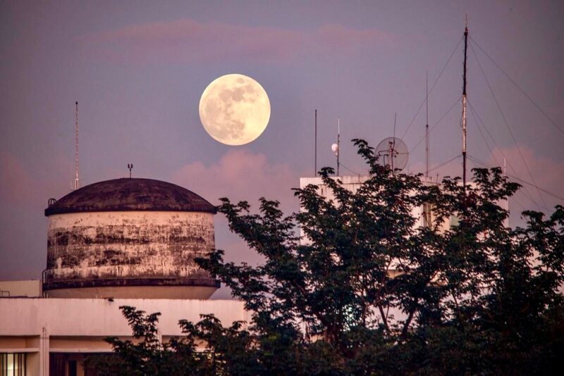 Large, white full moon over a rusty, dome-top water tower and a nearby tree.