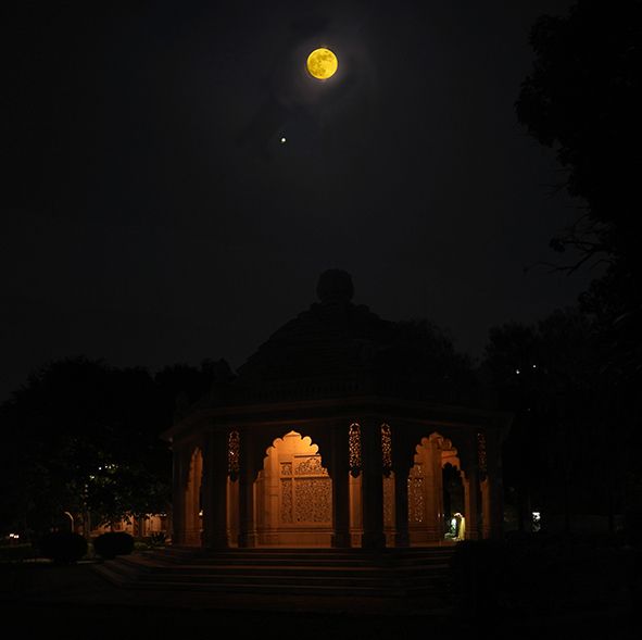 A darkened temple with a lighted doorway and above is a full moon and bright dot nearby that is Jupiter.
