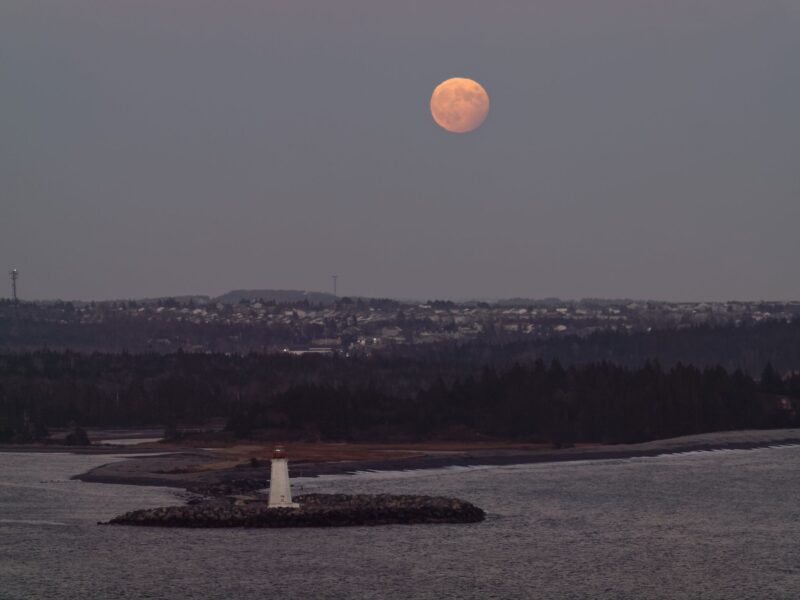 A reddish nearly full moon with a dark corner at lower right and a lighthouse next to the coast far below.