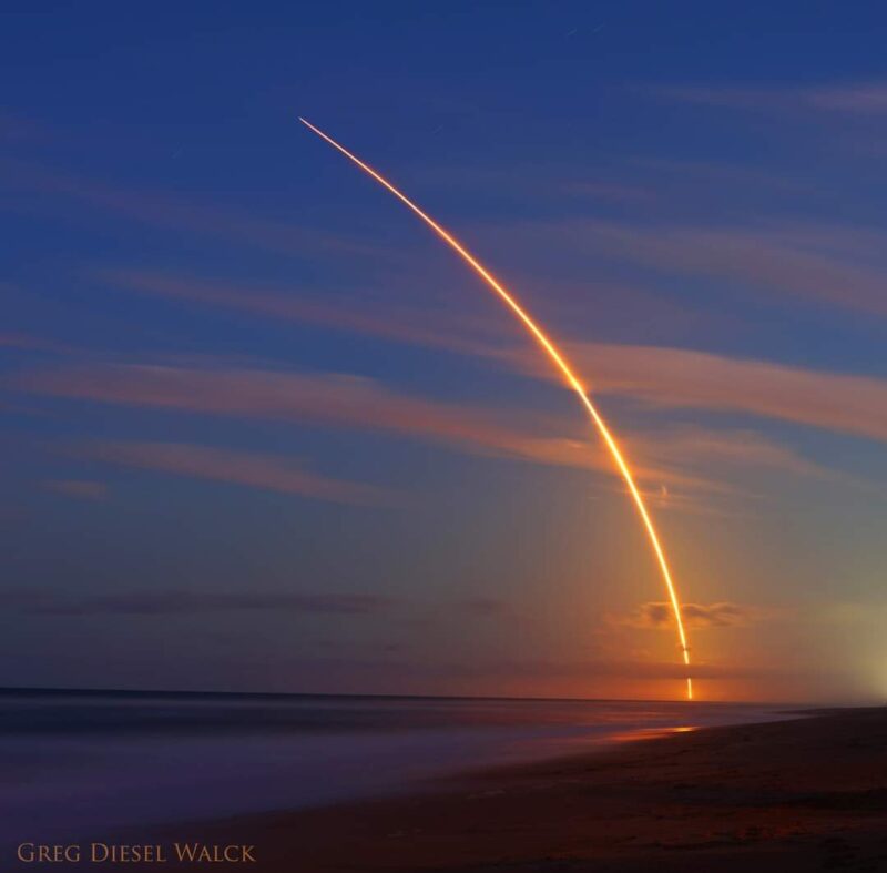 Illuminated arc of a launching rocket, over the ocean, at dusk.