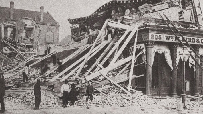 Black-and-white photo showing collapsed one-story city buildings with men standing around.