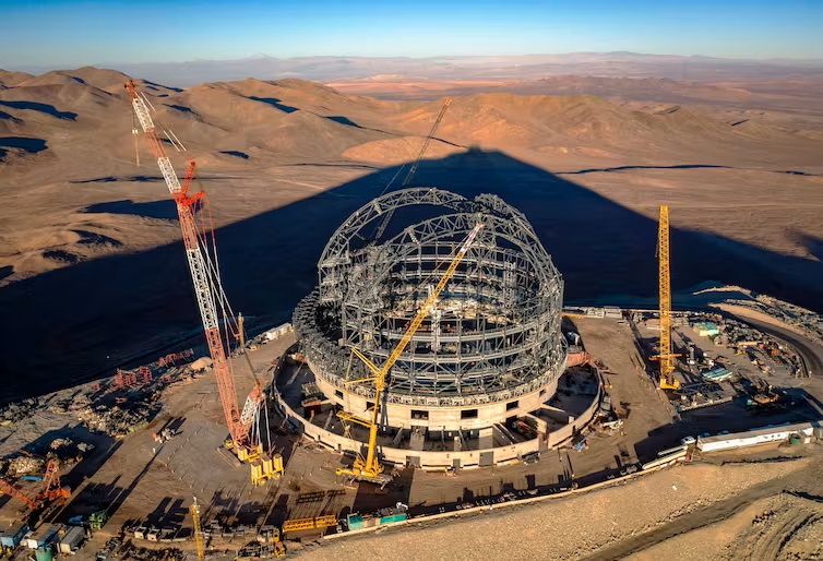 World's largest optical telescope: A construction site in a hilly desert showing the bracing for a round dome with cranes on either side.