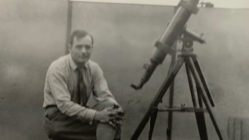 Black-and-white photo of a young man sitting next to a small telescope.