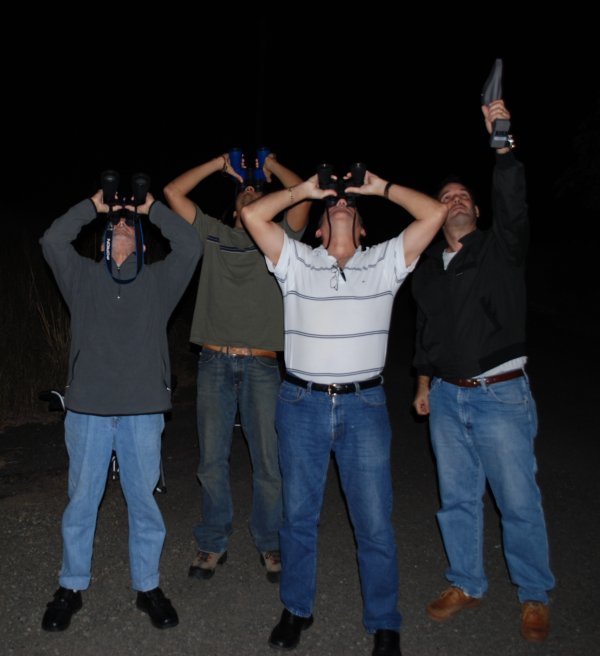 Four men outside under black sky looking skyward with binoculars.