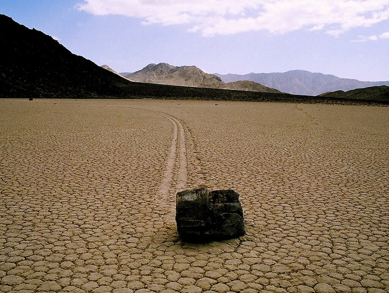 Sailing stone: Large squarish stone on dry, cracked desert ground with streaks behind it showing its path along the ground.