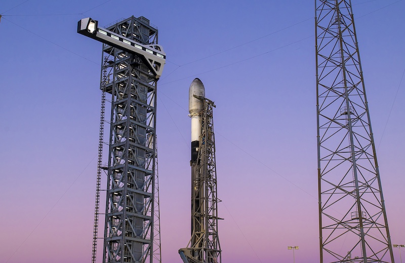 Starlink: A tall, narrow black and white rocket flanked by towers stands vertically against a pastel sunset sky.