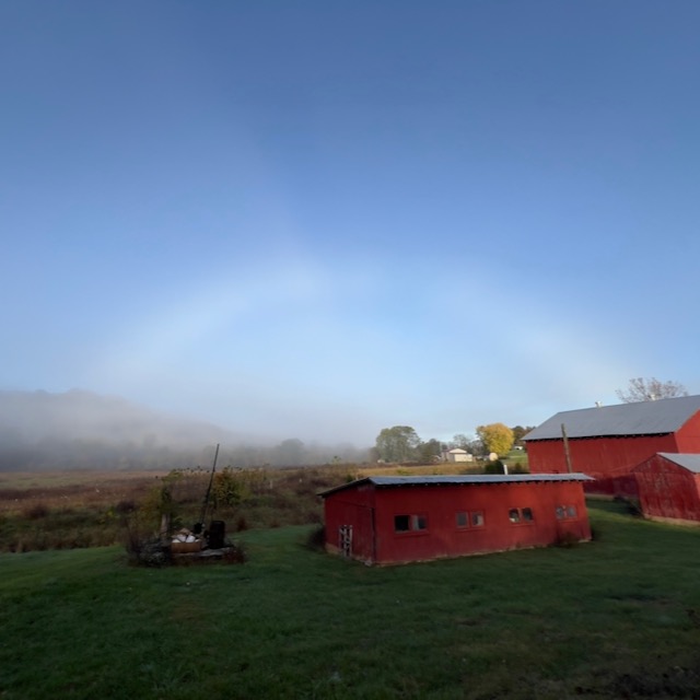 A fuzzy white arc in a blue sky above a bucolic scene with a red barn and a shed.