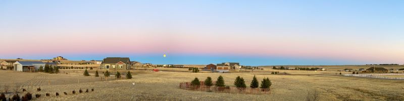 A panorama of a flat countryside with a few houses and on the far horizon is a curved band of blue with pink above.