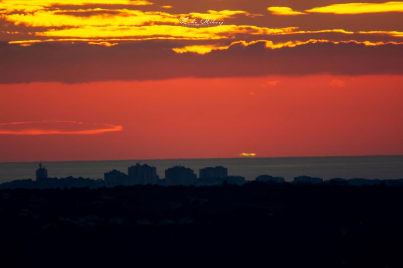 A view over a city and past a watery horizon to where a small line of yellowish green is surrounded by an orange sunset.