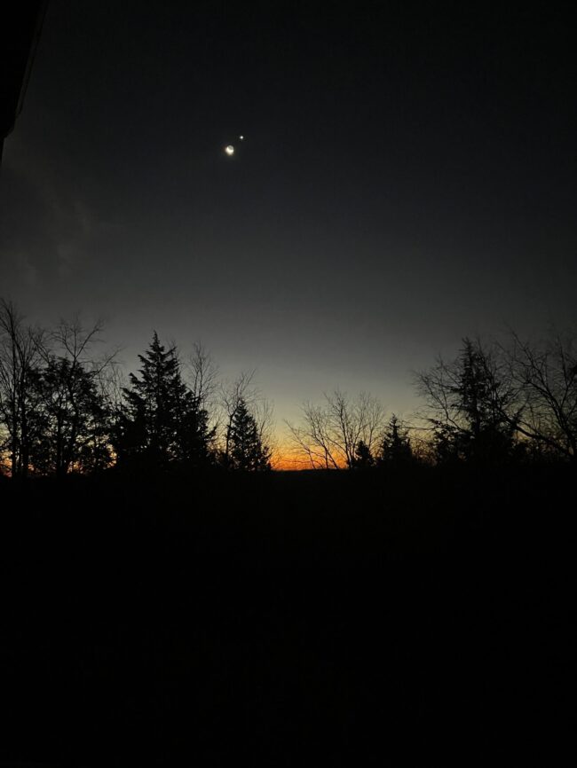 A dark scene with a rosy glow on the horizon and a crescent moon above near a bright dot.