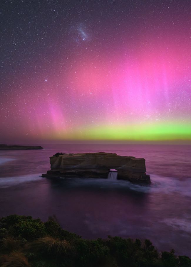 Pink and green light below small fuzzy glowing patch in sky and above rocky shoreline with a rock arch.