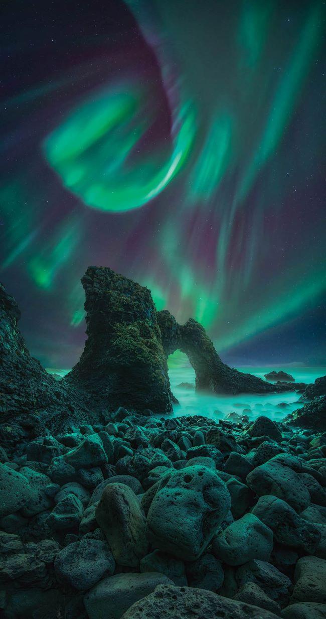Green curls of light in a night sky over a rocky scene with a rock arch over distant water.