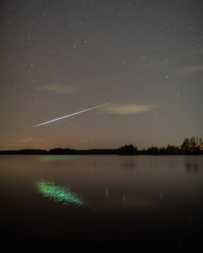 Starry sky with a white, long streak crossing it. It is reflected in the water in a green color.