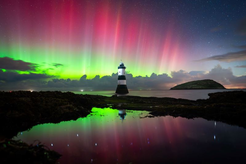 Northern lights photos: A lighthouse along a shoreline under vertical red and green streaks above distant clouds, the aurora reflected on the water.