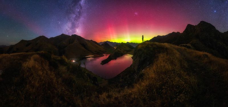 Red and yellow glowing arc of light above mountainous horizon with the Milky Way and a person in silhouette.