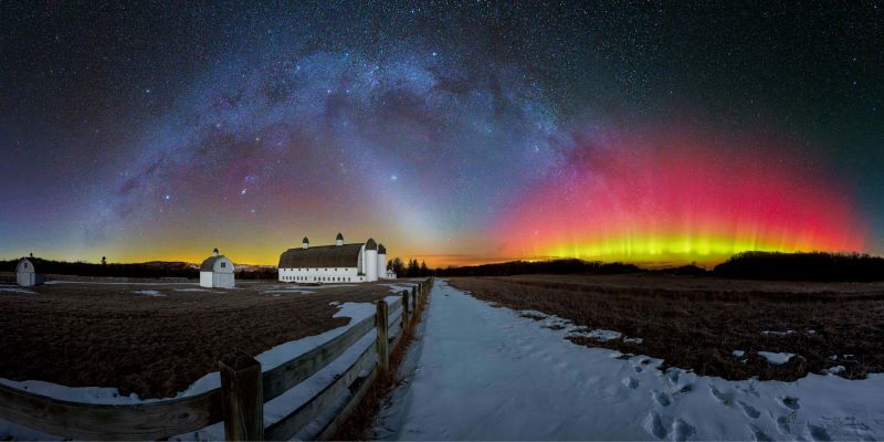 Arch of Milky Way and pink and yellow and auroral beams at horizon with a barn and a snowy road in the foreground.