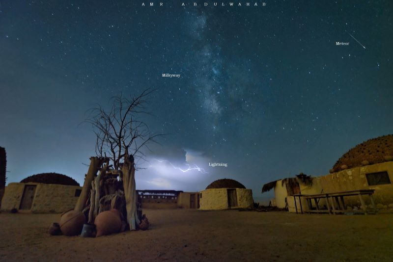 A desert landscape with huts and the Milky Way high above and lightning near the horizon. On the right is the streak of a meteor.