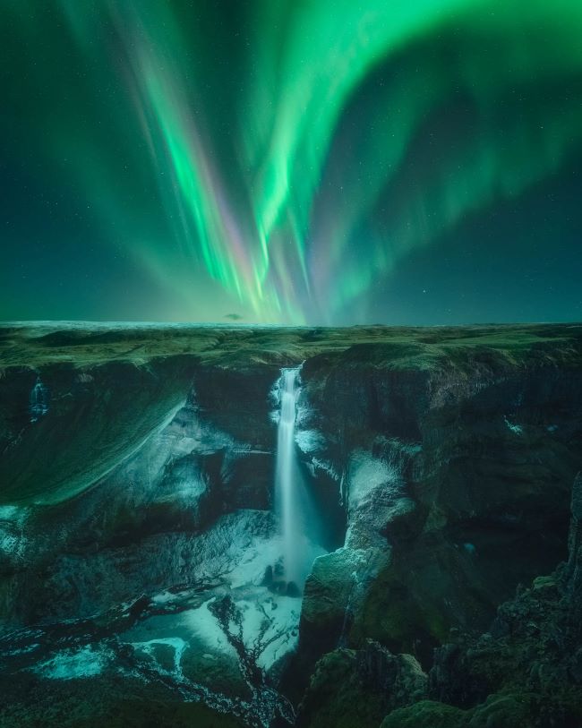 Several vertical curtain-like green streaks in night sky over a landscape with a tall, narrow waterfall in the distance.