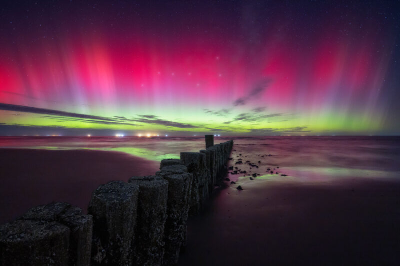 Band of vertical red and green streamers on the horizon glinting off water with log breakwater in the foreground.