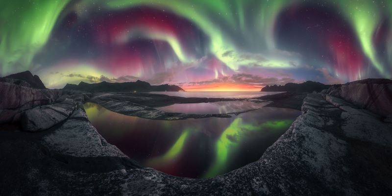 Green auroral streamers tinged with red in a fish-eye view over rocky landscape and large ponds of water.