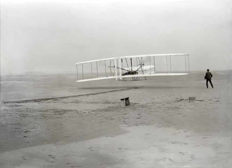 A flimsy-looking biplane in flight about a yard above flat sandy ground, with a man running beside it.