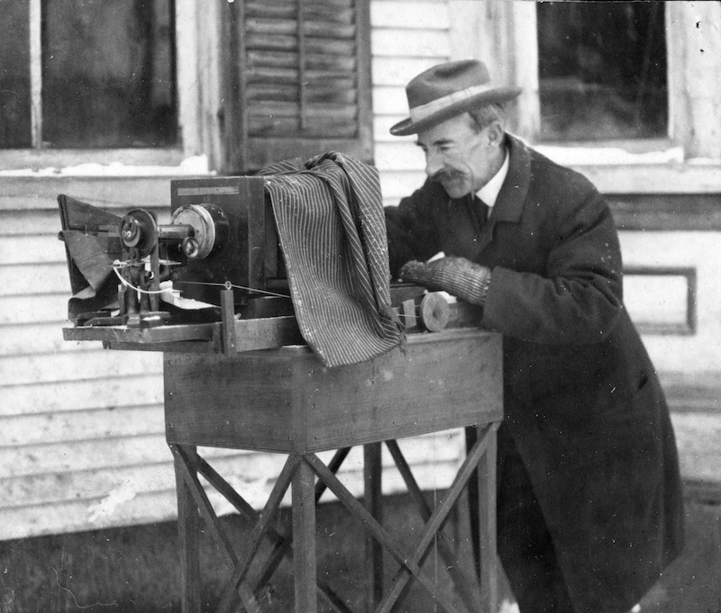A man in a winter coat and a hat standing in front of a large, old-fashioned camera setup on a table outside.
