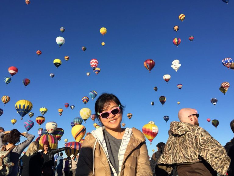 Woman with black hair and sunglasses looking at camera while many hot air balloons fill the sky behind.
