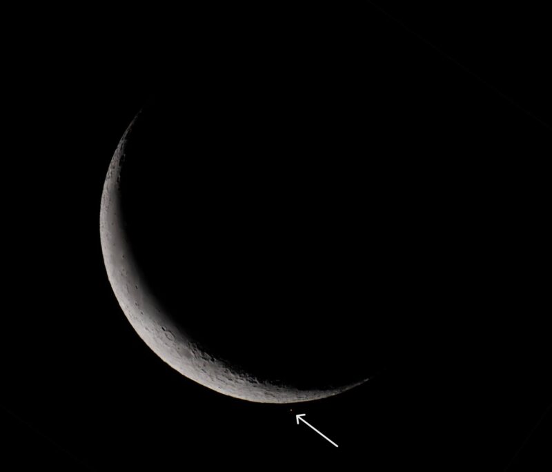 Slender crescent moon up close with a reddish dot just below it, that an arrow is pointing at.
