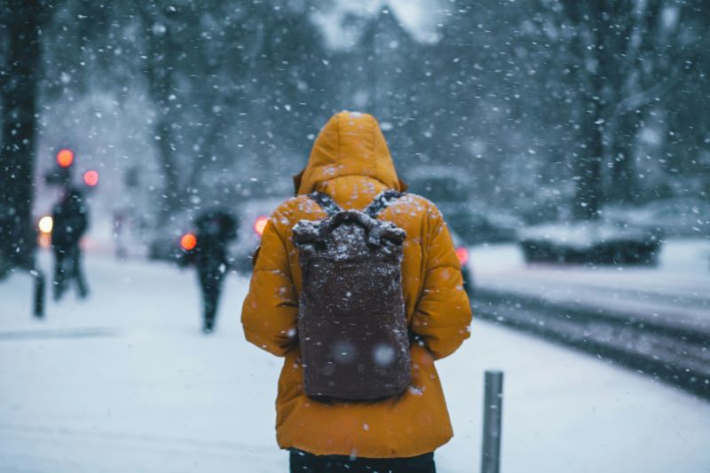 A person in a yellow coat with backpack walks through a snowy city scene.