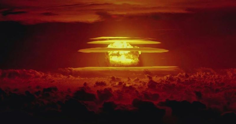 Underground nuclear tests: A distant, giant yellow fireball cloud with horizontal rings of clouds it, in a deep red sky.