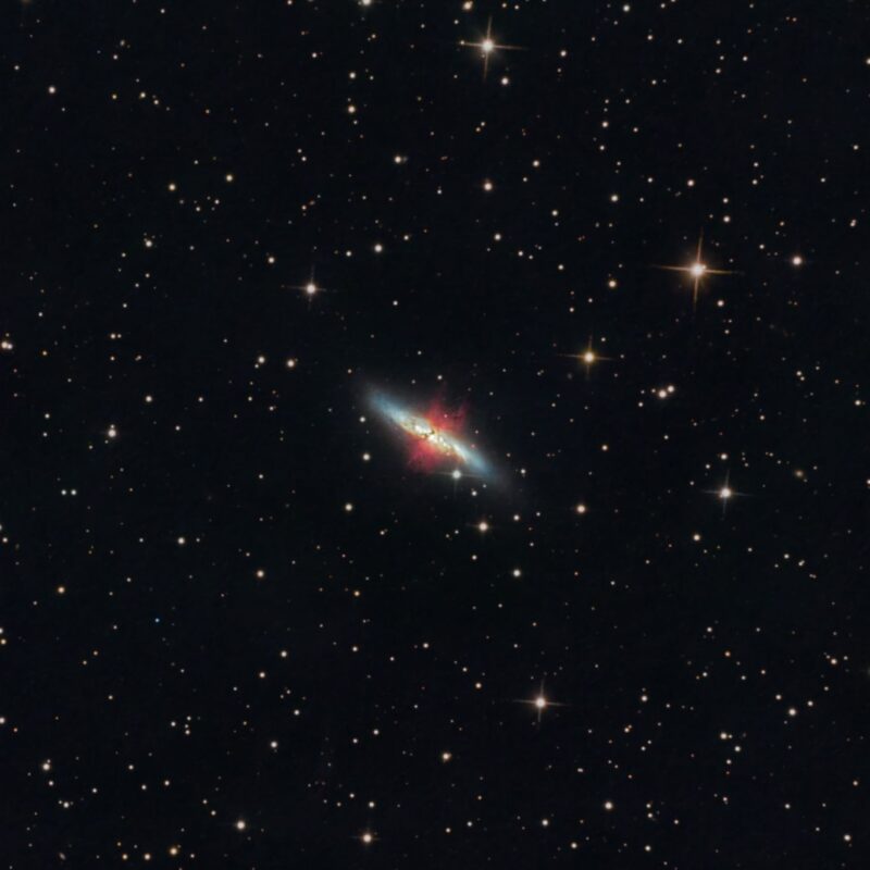 Large, cigar-shaped whitish nebula with a reddish burst and numerous foreground stars.