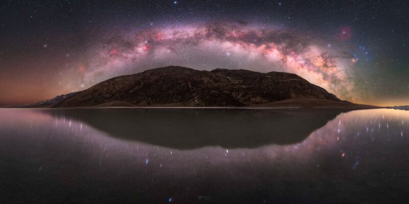 Milky Way arching over a dark mountain with a reflection in the water below.