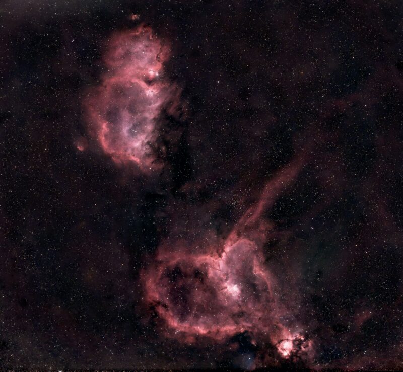 Two clouds of bright reddish nebulosity over a background of distant stars.